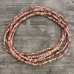 May include: A collection of beaded waist beads in shades of pink, rose, and gold. The beads are small and strung together, creating a delicate and elegant look. The beads are arranged in a circular pattern.