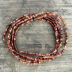 May include: A collection of beaded jewelry, featuring multiple strands of small, colorful beads. The beads are primarily in shades of copper, bronze, and purple, creating a warm, earthy color palette. The strands are arranged in a circular pattern.