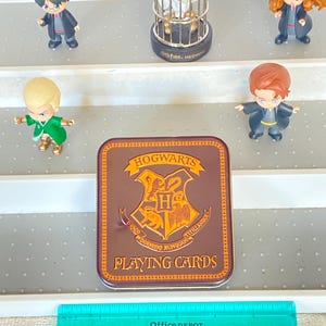 May include: A brown rectangular tin of Hogwarts playing cards with the Hogwarts crest. The tin is surrounded by small figurines of characters and a small cage with a bird inside. A green ruler is at the bottom.