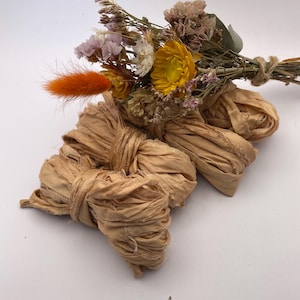 May include: A bundle of dried flowers in shades of yellow, pink, and white, with an orange accent, tied with twine. Below, several bundles of tan-colored, textured ribbon are arranged.