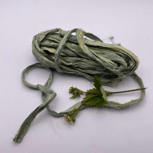 May include: A bundle of sage green ribbon is coiled on a white surface. A sprig of greenery with small white flowers and green leaves is next to the ribbon. The ribbon appears to be made of a soft, textured material.