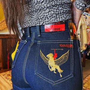 May include: Dark blue denim jeans with a decorative embroidered design on the back pocket. The design features a gold winged horse with the word "Devils" in red script above. The jeans have a classic five-pocket design and a straight leg.