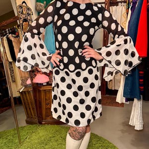 May include: A black and white polka dot dress with bell sleeves. The dress is paired with a black hat and white platform boots. The dress is knee-length and has a round neckline. The background includes clothing racks and a green rug.