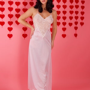 May include: A woman wearing a white lace slip dress with a sweetheart neckline, standing in front of a pink wall with red heart decorations.