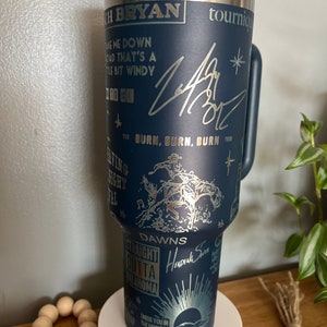 May include: A blue stainless steel tumbler with a gold design featuring the text "Take me down a road that's a little bit windy", "Burn, Burn, Burn Tour", "Dawns", "Straight outta Oklahoma", and "I miss you in the morning when I see the sun". The tumbler also features a gold signature and a gold star.