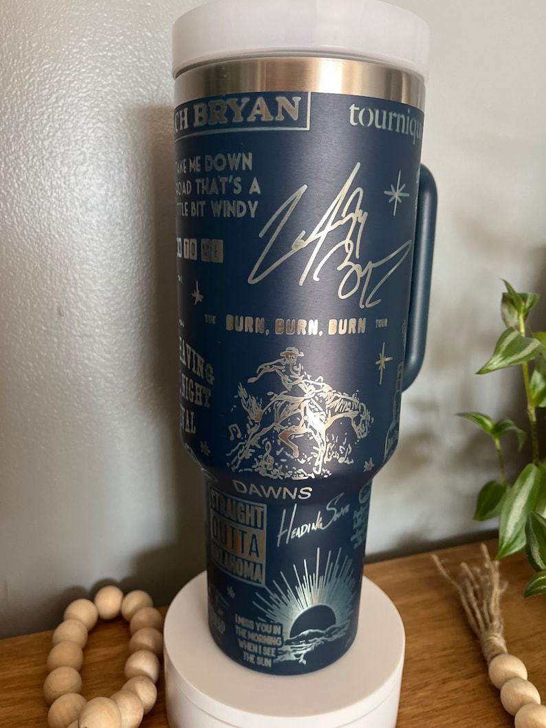 May include: A blue stainless steel tumbler with a gold design featuring the text "Take me down a road that's a little bit windy", "Burn, Burn, Burn Tour", "Dawns", "Straight outta Oklahoma", and "I miss you in the morning when I see the sun". The tumbler also features a gold signature and a gold star.