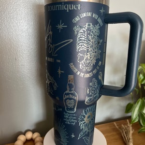 May include: A navy blue stainless steel travel mug with a black handle. The mug has a variety of silver metallic designs including a skeleton, flowers, a bottle of Jim Beam, a sunflower, a nautical anchor, and the text "tourniquet", "Find someone who grows flowers in the darkest part of you", "sweetest of the", and "Baptize me in a bottle of tears and put Johnny Cash on the vinyl".