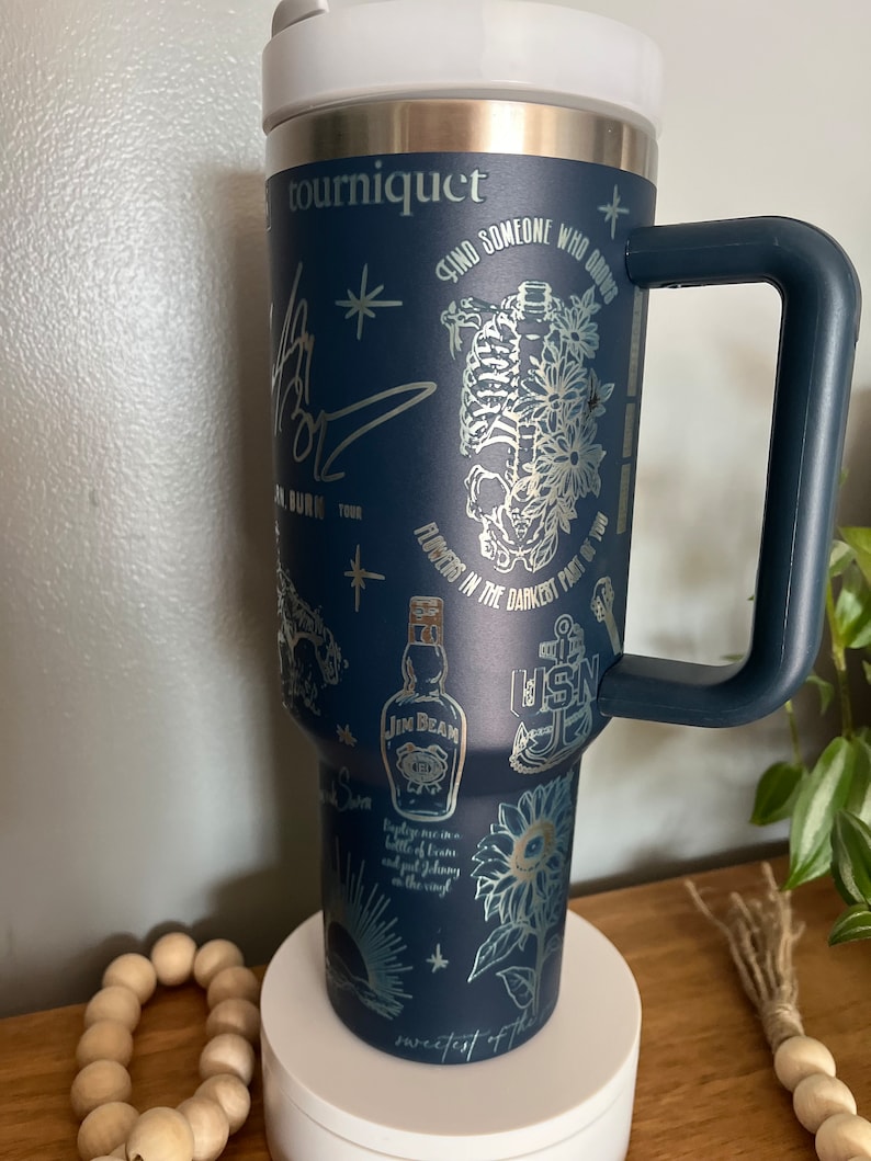 May include: A navy blue stainless steel travel mug with a black handle. The mug has a variety of silver metallic designs including a skeleton, flowers, a bottle of Jim Beam, a sunflower, a nautical anchor, and the text "tourniquet", "Find someone who grows flowers in the darkest part of you", "sweetest of the", and "Baptize me in a bottle of tears and put Johnny Cash on the vinyl".