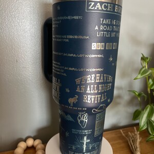 May include: A blue stainless steel tumbler with a silver design featuring a cowboy hat, a heart, a cross, and the text "Zach Bryan, We're Having An All Night Revival, Sun to Me, Give Your Heart Hit Keep Your Head Up, The Devil Can Scrap But The Lord Harms".