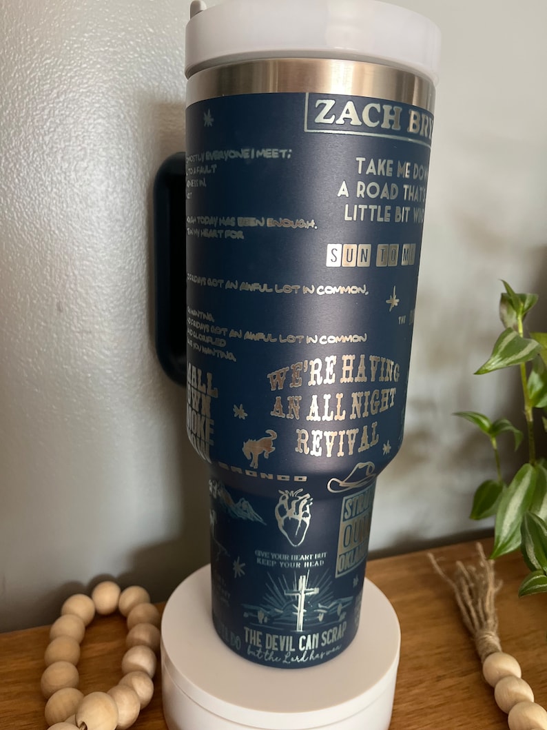 May include: A blue stainless steel tumbler with a silver design featuring a cowboy hat, a heart, a cross, and the text "Zach Bryan, We're Having An All Night Revival, Sun to Me, Give Your Heart Hit Keep Your Head Up, The Devil Can Scrap But The Lord Harms".
