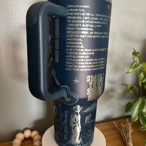 May include: A blue stainless steel tumbler with a black handle. The tumbler has a gold design with a quote and a drawing of a cowboy. The text on the tumbler reads "Small Town Smoke Show".
