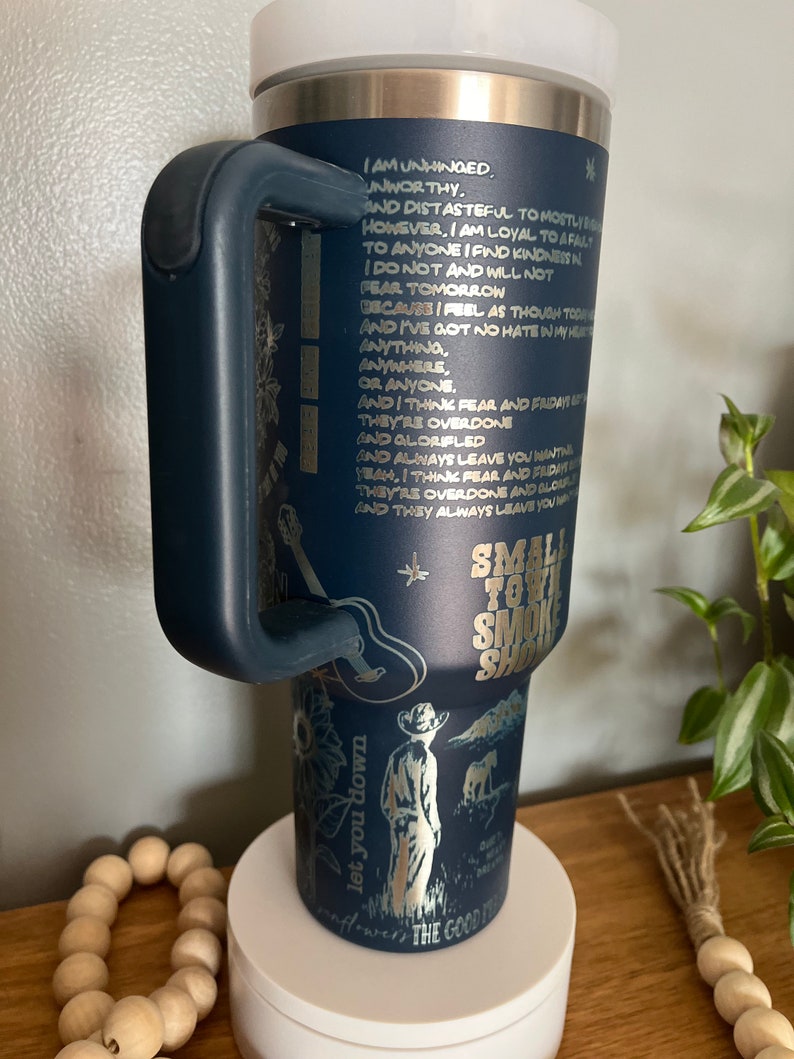 May include: A blue stainless steel tumbler with a black handle. The tumbler has a gold design with a quote and a drawing of a cowboy. The text on the tumbler reads "Small Town Smoke Show".