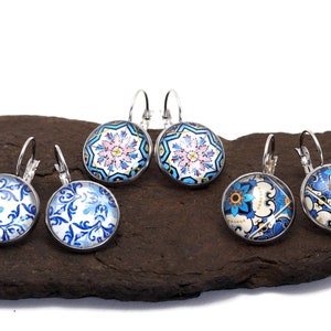 Peut inclure: Six paires de boucles d'oreilles rondes à fermoir à levier argentées, avec des motifs colorés. Les motifs comprennent des motifs floraux et géométriques dans les tons de bleu, blanc, rose et jaune. Les boucles sont présentées sur une surface sombre et texturée.