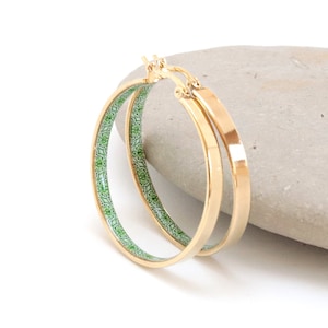 Könnte beinhalten: Goldfarbene Creolen. Die Ohrringe haben eine flache, polierte goldene Außenseite und eine grün-weiß gemusterte Innenseite. Die Ohrringe werden auf einem grauen Stein vor weißem Hintergrund präsentiert. Ein klassisches Schmuckaccessoire.