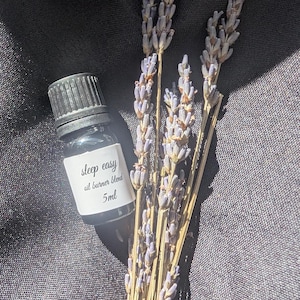 May include: A small, dark glass bottle with a black cap and a white label that reads "sleep easy oil burner blend 5ml" is next to dried lavender stems. The lavender has purple buds and brown stems. The background is a dark, textured fabric.