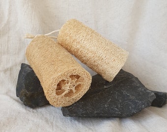 May include: Two natural loofah sponges, cylindrical in shape, are displayed on dark grey stones against a neutral fabric background. The sponges are a light beige colour, with a textured, porous surface. One sponge has a string loop.