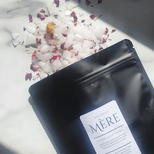 May include: A black pouch with a white label that reads "Petit Papillon Mère Sootheing & Rejuvenating". The label lists the ingredients and directions for use. The pouch is open and white bath salts with dried flowers are spilling out.