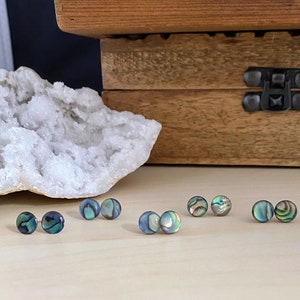 May include: A set of six iridescent abalone shell earrings. The earrings are round and have a blue, green, and gold color scheme.