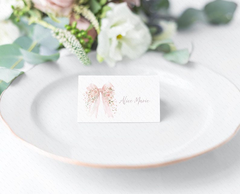 Pode incluir: Um cart&atilde;o de lugar branco com um la&ccedil;o e um design floral em aquarela. O cart&atilde;o tem o nome "Alice Marie" escrito em it&aacute;lico. Flores rosa e verde suaves no fundo, com um prato branco.