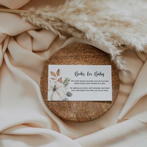 May include: A white card with the text "Books for Baby" sits on a wooden plate. The card features a watercolor pumpkin and fall foliage. The card's text requests guests bring a book instead of a card.