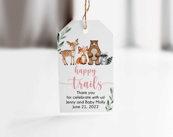 May include: A white gift tag with a watercolor illustration of woodland animals, including a deer, fox, bear, and raccoon. The tag reads "happy trails" in pink script, with additional text below. The tag is tied with a brown string.