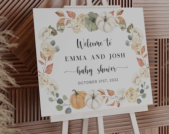 May include: A white sign on a wooden easel with the text "Welcome to EMMA AND JOSH baby shower OCTOBER 21ST, 2022". The sign is decorated with a floral wreath of pumpkins, roses, and leaves in shades of orange, green, and cream.