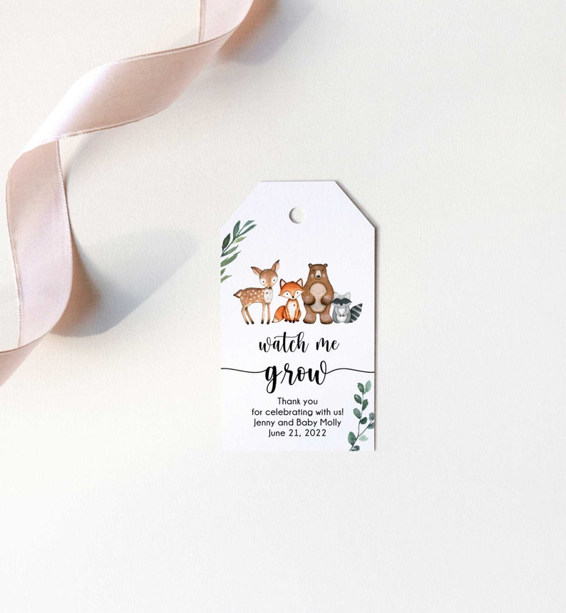 May include: A white tag with a green leafy border and a watercolor illustration of a deer, fox, bear, and raccoon. The text "watch me grow" is written in black cursive font. The text "Thank you for celebrating with us! Jenny and Baby Molly June 21, 2022" is written in black font.