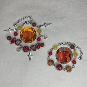 May include: Two silver charm bracelets with red, brown, and yellow beads. Each bracelet features a charm of a red apple with gold stars and a hand holding the apple.