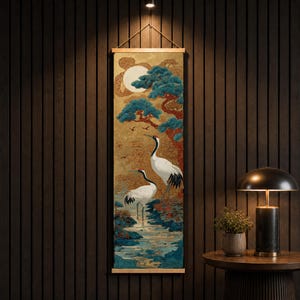 Könnte beinhalten: Ein vertikaler Wandbehang mit einer detaillierten Illustration von zwei weißen Kranichen mit schwarz gefiederten Flügeln, die in einem blauen Bach stehen. Der Hintergrund zeigt einen goldenen Himmel, einen großen weißen Mond und stilisierte blaue Kiefern.