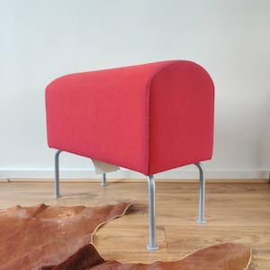 May include: A red upholstered bench with a curved back and four silver metal legs. The bench is set on a wooden floor, with a brown rug in the foreground. The bench is a modern furniture piece.
