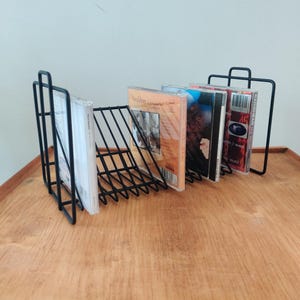 May include: A black metal CD holder with a handle, holding several CDs. The holder has a wireframe design and is sitting on a wooden surface. The CDs have various album covers.