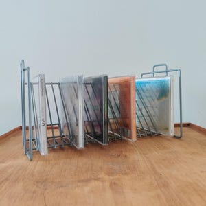 May include: A silver metal CD rack holding several CD cases. The rack has a wireframe design and is sitting on a wooden surface. The CDs are visible, with various album art displayed.