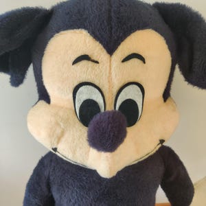 K&ouml;nnte beinhalten: Vintage Mickey Mouse Pl&uuml;schtier. Das Pl&uuml;schtier hat einen gro&szlig;en Kopf mit schwarzen Ohren, einem cremefarbenen Gesicht und einer lila Nase. Der K&ouml;rper ist marineblau mit wei&szlig;en Handschuhen. Das Spielzeug hat einen l&auml;chelnden Ausdruck.