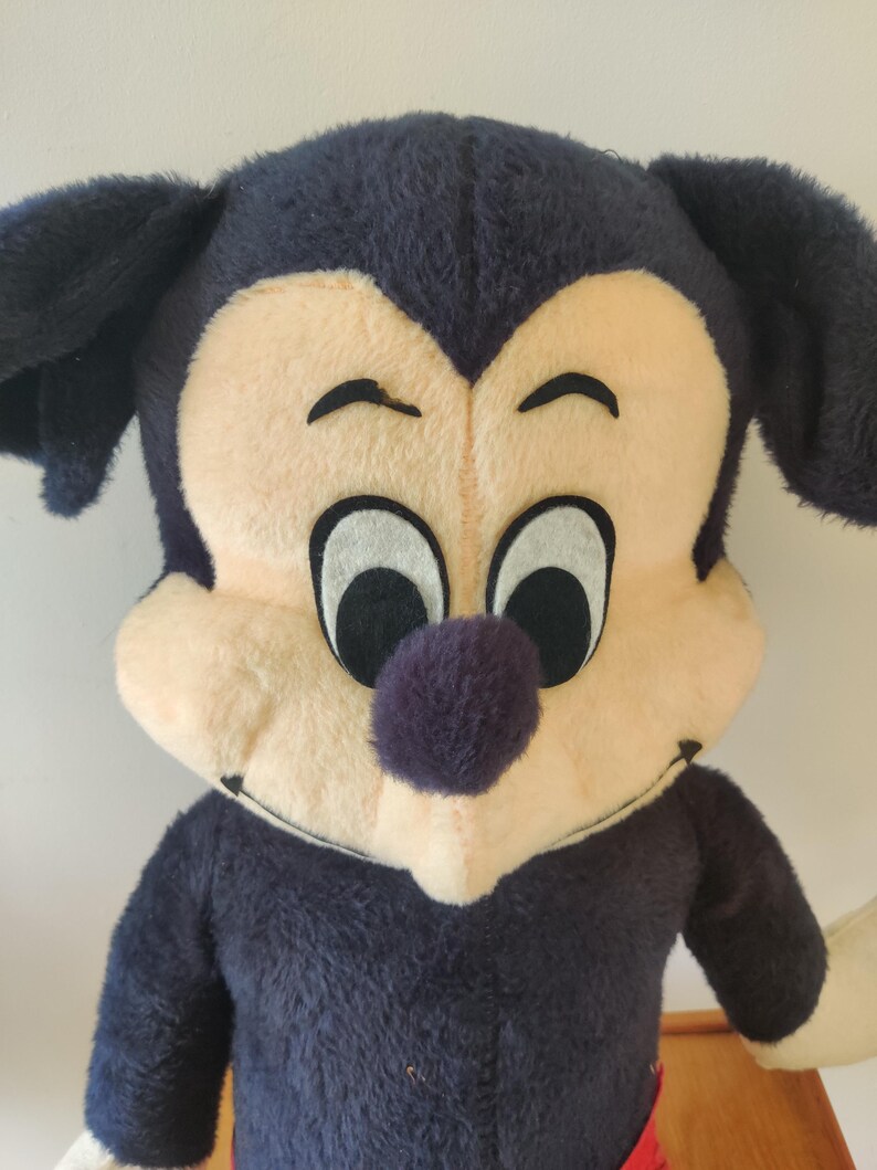 K&ouml;nnte beinhalten: Vintage Mickey Mouse Pl&uuml;schtier. Das Pl&uuml;schtier hat einen gro&szlig;en Kopf mit schwarzen Ohren, einem cremefarbenen Gesicht und einer lila Nase. Der K&ouml;rper ist marineblau mit wei&szlig;en Handschuhen. Das Spielzeug hat einen l&auml;chelnden Ausdruck.