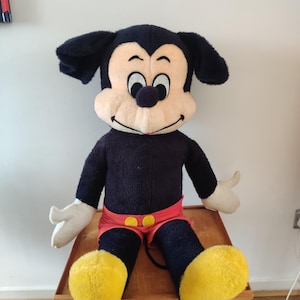K&ouml;nnte beinhalten: Eine gro&szlig;e Mickey Mouse Pl&uuml;schpuppe. Die Puppe hat einen schwarzen K&ouml;rper, Ohren und Beine, mit einem cremefarbenen Gesicht und H&auml;nden. Mickey tr&auml;gt rote Shorts mit gelben Kn&ouml;pfen und gro&szlig;e gelbe Schuhe. Die Puppe sitzt auf einem Holztisch.