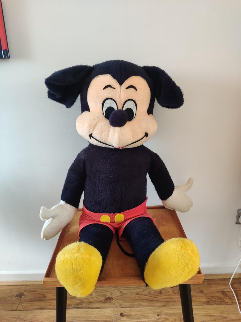 K&ouml;nnte beinhalten: Eine gro&szlig;e Mickey Mouse Pl&uuml;schpuppe. Die Puppe hat einen schwarzen K&ouml;rper, Ohren und Beine, mit einem cremefarbenen Gesicht und H&auml;nden. Mickey tr&auml;gt rote Shorts mit gelben Kn&ouml;pfen und gro&szlig;e gelbe Schuhe. Die Puppe sitzt auf einem Holztisch.