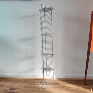 May include: A tall, silver metal CD rack with four square shelves. The rack is made of thin, vertical wires and has a rectangular base. The shelves are made of a light-colored material. The rack is standing on a wooden floor.