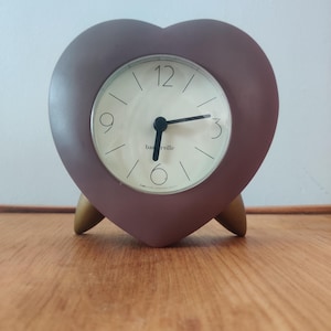 Puede incluir: Un reloj en forma de corazón con un marco malva y una esfera crema. El reloj tiene manecillas negras y un diseño minimalista, apoyado en tres patas de madera. La esfera del reloj muestra la palabra "basrville".