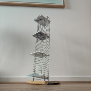 May include: A tall, vertical chrome wire storage rack designed for compact discs, featuring a rectangular wooden base. The metal frame consists of multiple horizontal slots for organizing individual cases, with four CD cases currently inserted.