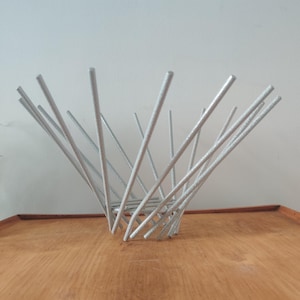 May include: A silver metal decorative bowl with a spiky design. The bowl is made of thin, straight rods that radiate outwards from a central base. The bowl is sitting on a wooden surface.