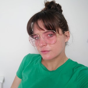 May include: A woman with brown hair wearing a green shirt and gold-rimmed glasses with pink lenses.