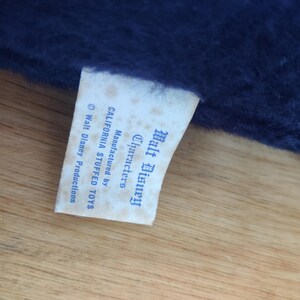 K&ouml;nnte beinhalten: Nahaufnahme eines Etiketts von einem Walt Disney Stofftier. Das cremefarbene Etikett zeigt blauen Text, darunter "Walt Disney Characters" und "Manufactured by CALIFORNIA STUFFED TOYS". Das Etikett ist an einem dunkelblauen Pl&uuml;schstoff befestigt.