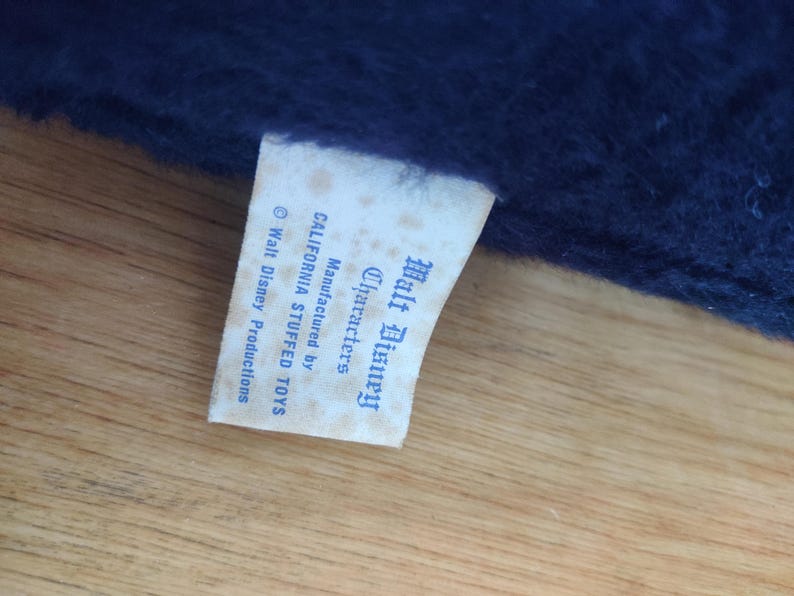 K&ouml;nnte beinhalten: Nahaufnahme eines Etiketts von einem Walt Disney Stofftier. Das cremefarbene Etikett zeigt blauen Text, darunter "Walt Disney Characters" und "Manufactured by CALIFORNIA STUFFED TOYS". Das Etikett ist an einem dunkelblauen Pl&uuml;schstoff befestigt.