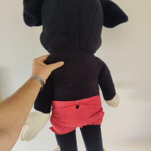 K&ouml;nnte beinhalten: Ein Vintage-Mickey-Mouse-Pl&uuml;schtier, von hinten gesehen. Das Pl&uuml;schtier hat einen schwarzen K&ouml;rper, gro&szlig;e Ohren, rote Shorts, wei&szlig;e Handschuhe und gelbe Schuhe. Das Spielzeug wird von der Hand einer Person gehalten.