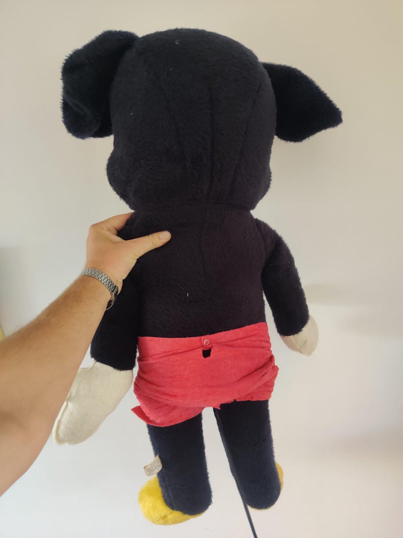 K&ouml;nnte beinhalten: Ein Vintage-Mickey-Mouse-Pl&uuml;schtier, von hinten gesehen. Das Pl&uuml;schtier hat einen schwarzen K&ouml;rper, gro&szlig;e Ohren, rote Shorts, wei&szlig;e Handschuhe und gelbe Schuhe. Das Spielzeug wird von der Hand einer Person gehalten.