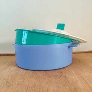 Könnte beinhalten: Ein Stapel Vintage-Tupperware-Behälter in Teal, Hellblau und Weiß. Das Set umfasst einen großen, runden, hellblauen Behälter, einen kleineren tealfarbenen Behälter und einen weißen Deckel mit einem tealfarbenen Griff. Die Behälter stehen auf einer Holzoberfläche.