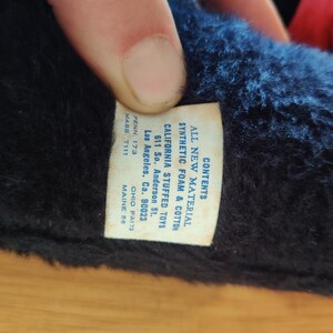 K&ouml;nnte beinhalten: Nahaufnahme eines Vintage-Stofftiers mit Etikett. Das Etikett lautet "ALL NEW MATERIAL SYNTHETIC FOAM & COTTON CALIFORNIA STUFFED TOYS" mit einer Adresse in Los Angeles, CA. Das Fell des Spielzeugs ist dunkelblau und schwarz.
