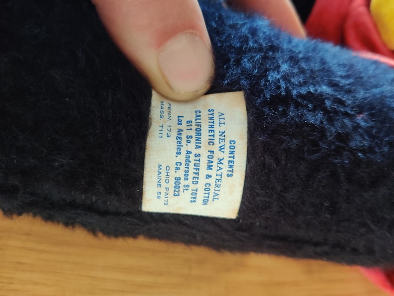 K&ouml;nnte beinhalten: Nahaufnahme eines Vintage-Stofftiers mit Etikett. Das Etikett lautet "ALL NEW MATERIAL SYNTHETIC FOAM & COTTON CALIFORNIA STUFFED TOYS" mit einer Adresse in Los Angeles, CA. Das Fell des Spielzeugs ist dunkelblau und schwarz.