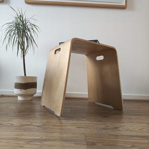 May include: A light-colored bent plywood stool featuring a smooth, curved top surface and two rectangular cut-out handles on the side panels. The stool has a simple, U-shaped structure with a natural wood grain finish and clean, rounded edges.