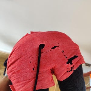 K&ouml;nnte beinhalten: Ein rotes, abgenutztes Stoffelement mit einer schwarzen Kordel, m&ouml;glicherweise Teil eines Kost&uuml;ms oder einer Requisite. Der Stoff weist mehrere L&ouml;cher und Risse auf, was auf Abnutzung hindeutet. Der Hintergrund ist hell und neutral.